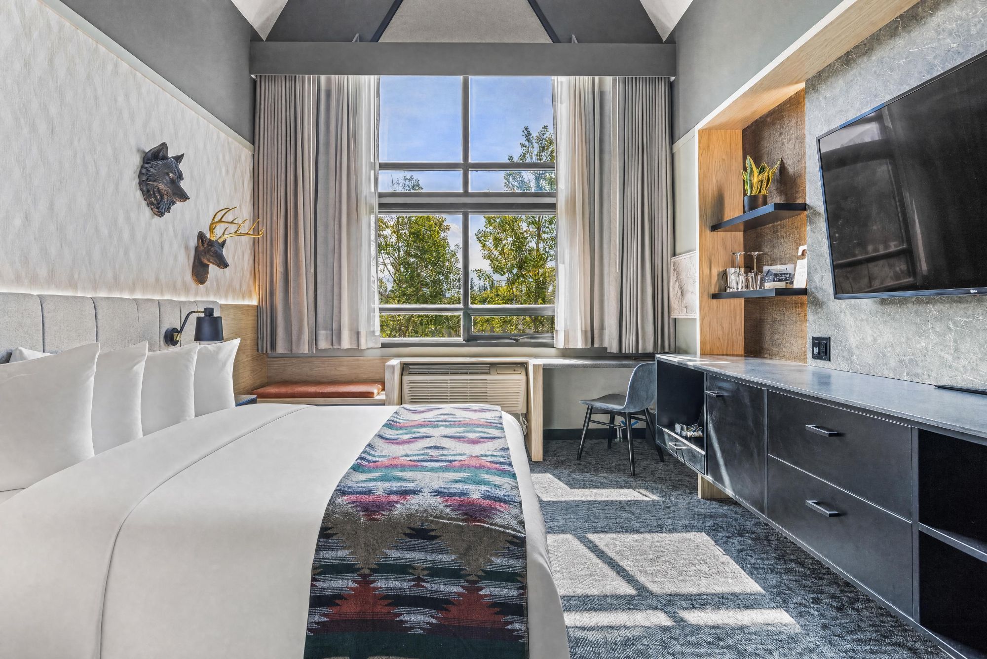 Una habitación de hotel moderna con una cama grande, un escritorio, una silla, un televisor y un ventanal con cortinas.