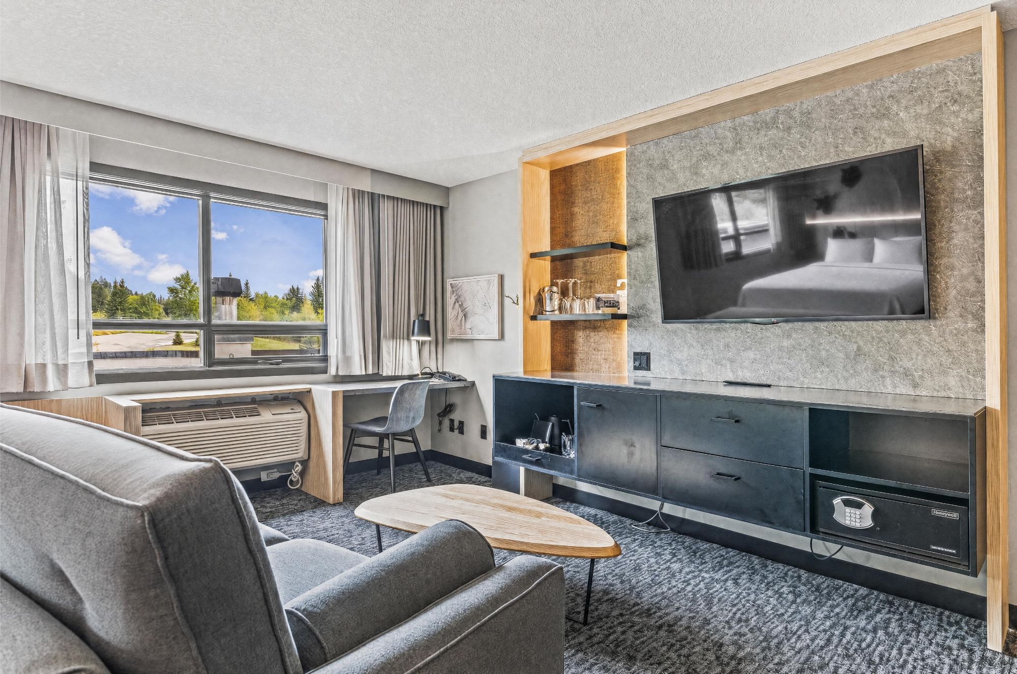 A modern hotel room with a large bed, a flat-screen TV mounted on a stone wall, a desk with a chair, and a sofa.