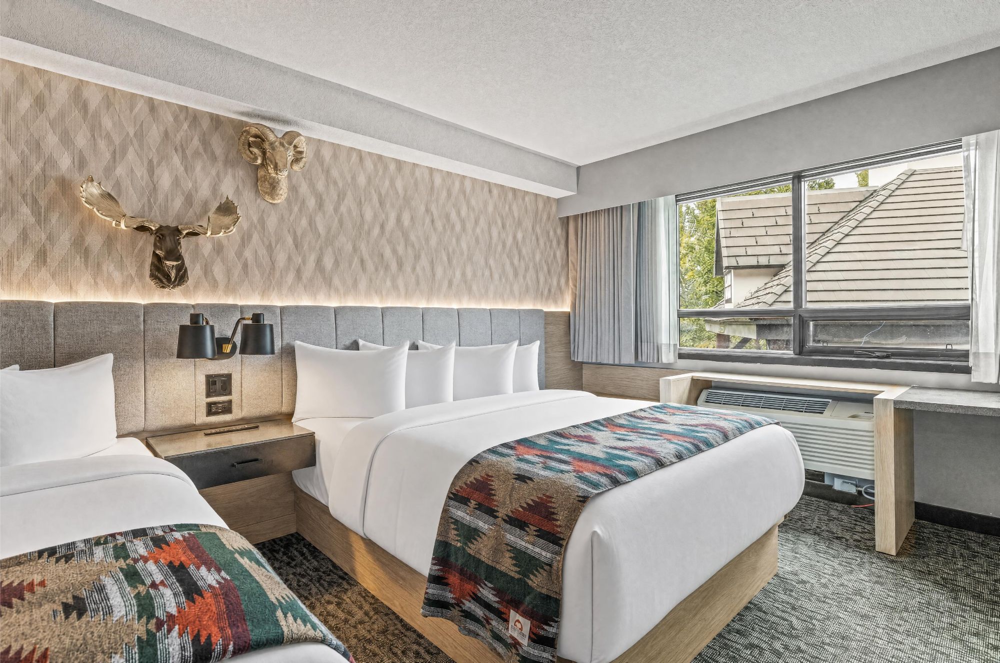 A hotel room with two beds, a desk, a mounted ram head decoration, and a window with a view of a building.