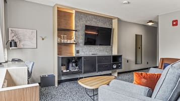 Una sala de estar moderna con un sofá gris, una mesa de centro de madera, un televisor de pantalla plana montado en una pared con textura, y un pequeño escritorio de madera con una lámpara.
