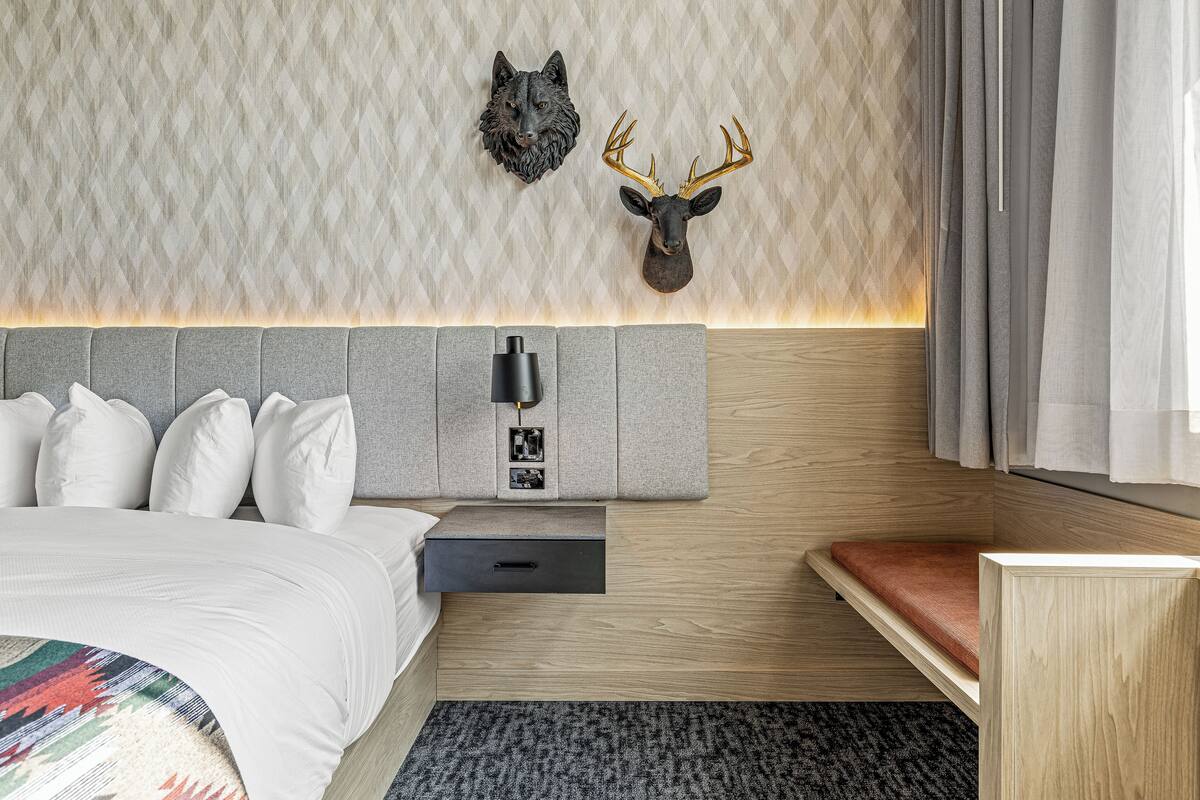 Une chambre d’hôtel moderne comprenant un lit, une table de chevet, une tête de lit en bois, une lampe fixée au mur et une décoration représentant une tête de cerf.
