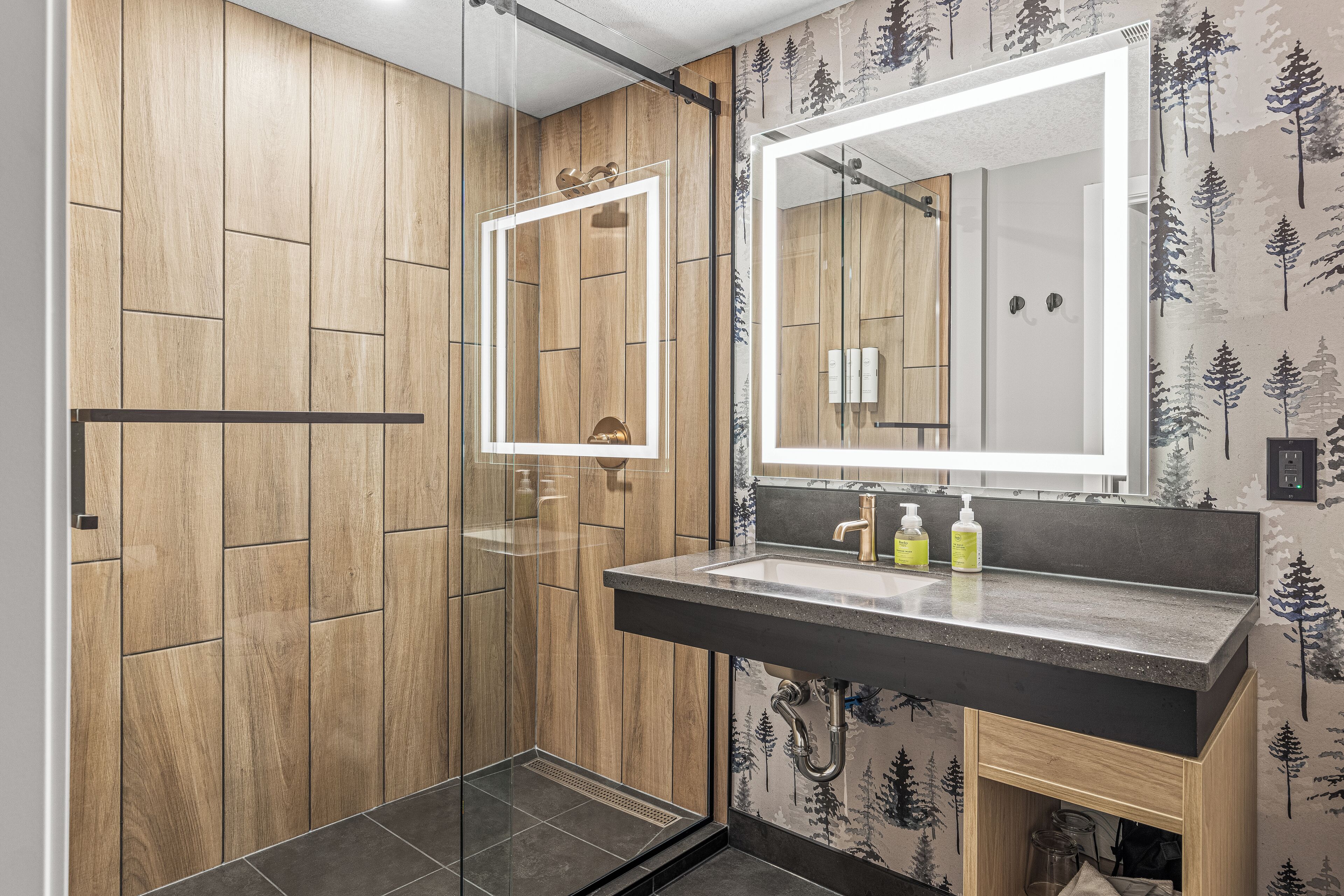 A modern bathroom with a wooden panel wall, a glass shower enclosure, a large mirror, and a black countertop with two bottles of soap.