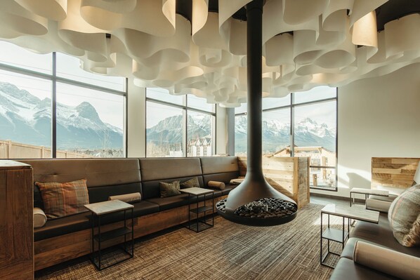 A modern interior with a sofa, tables, and a fireplace. Large windows offer mountain views. - Everwild Canmore Hotel (Canmore)