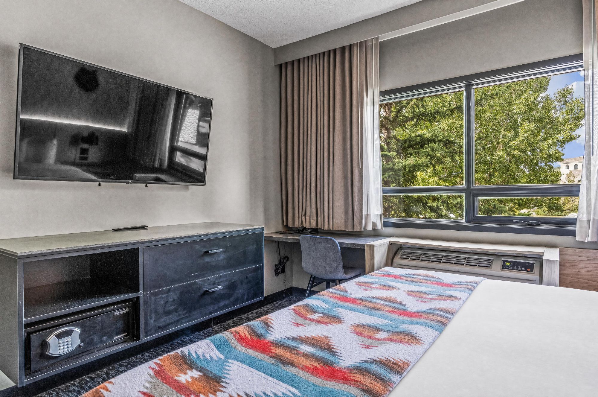 Une chambre d’hôtel dotée d’un téléviseur à écran plat fixé au mur, d’un bureau avec une chaise, d’une fenêtre avec des rideaux et d’un tapis à motifs.