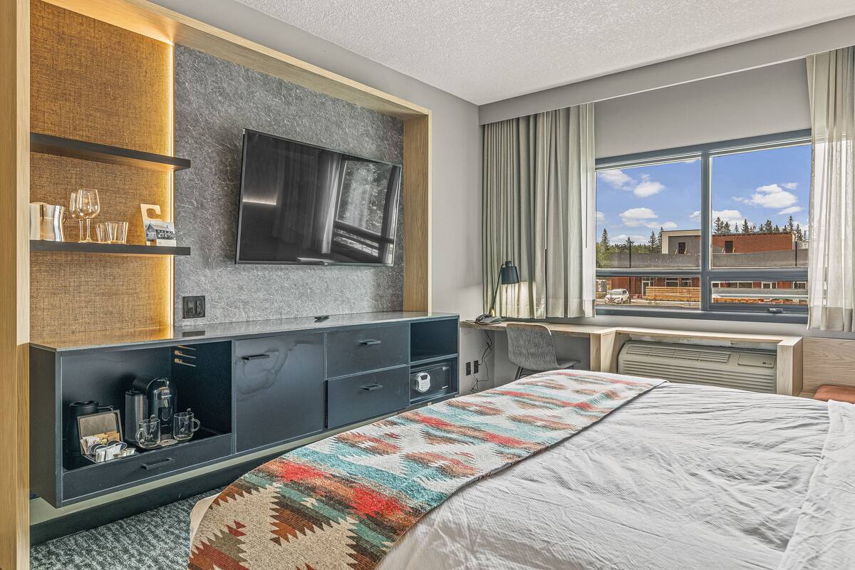 Une chambre d’hôtel avec un lit, un téléviseur fixé au mur, un bureau avec une chaise et une fenêtre donnant sur des immeubles.