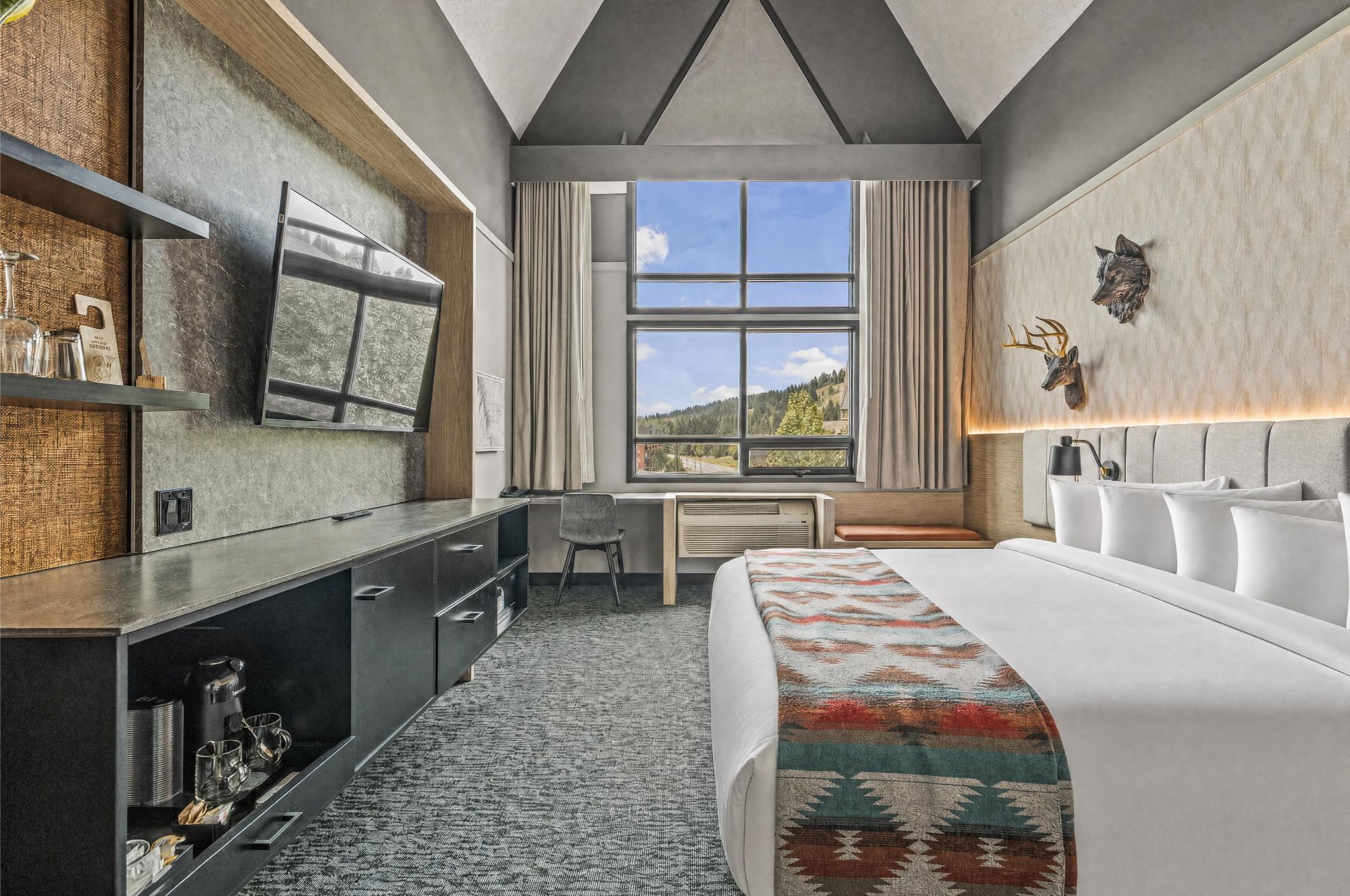 A modern hotel room with a large bed, a desk, a chair, and a window with a view of the outdoors.