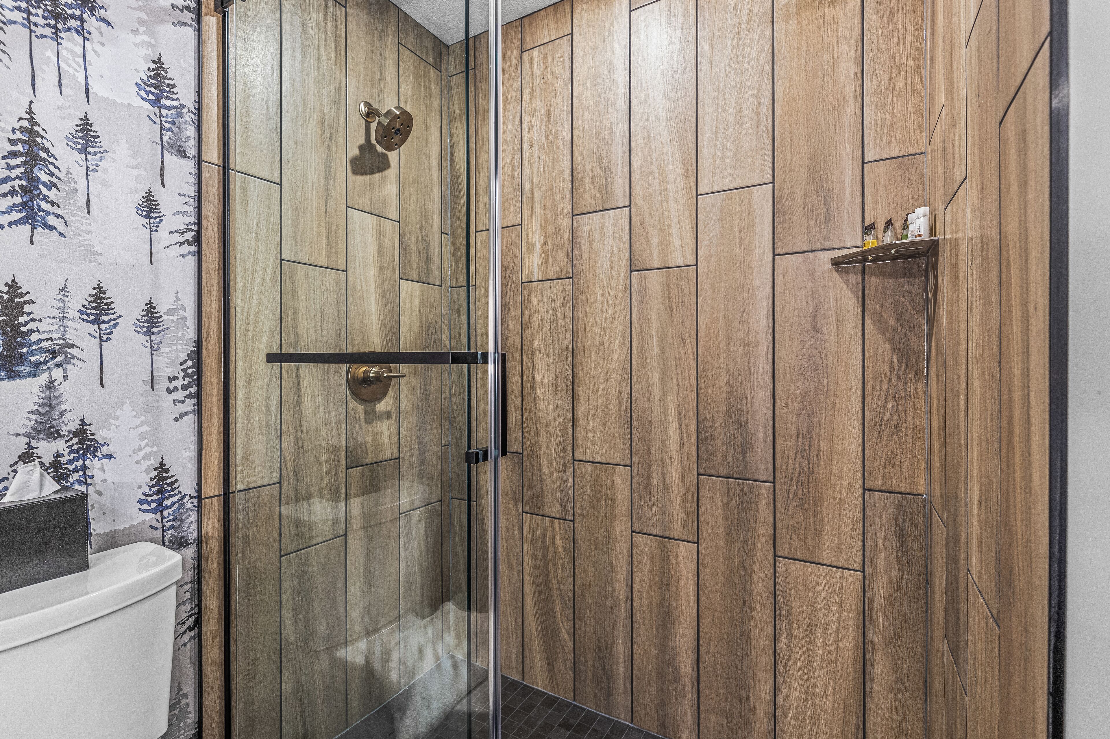 A shower with a glass door, wooden panel walls, and a shelf with toiletries.