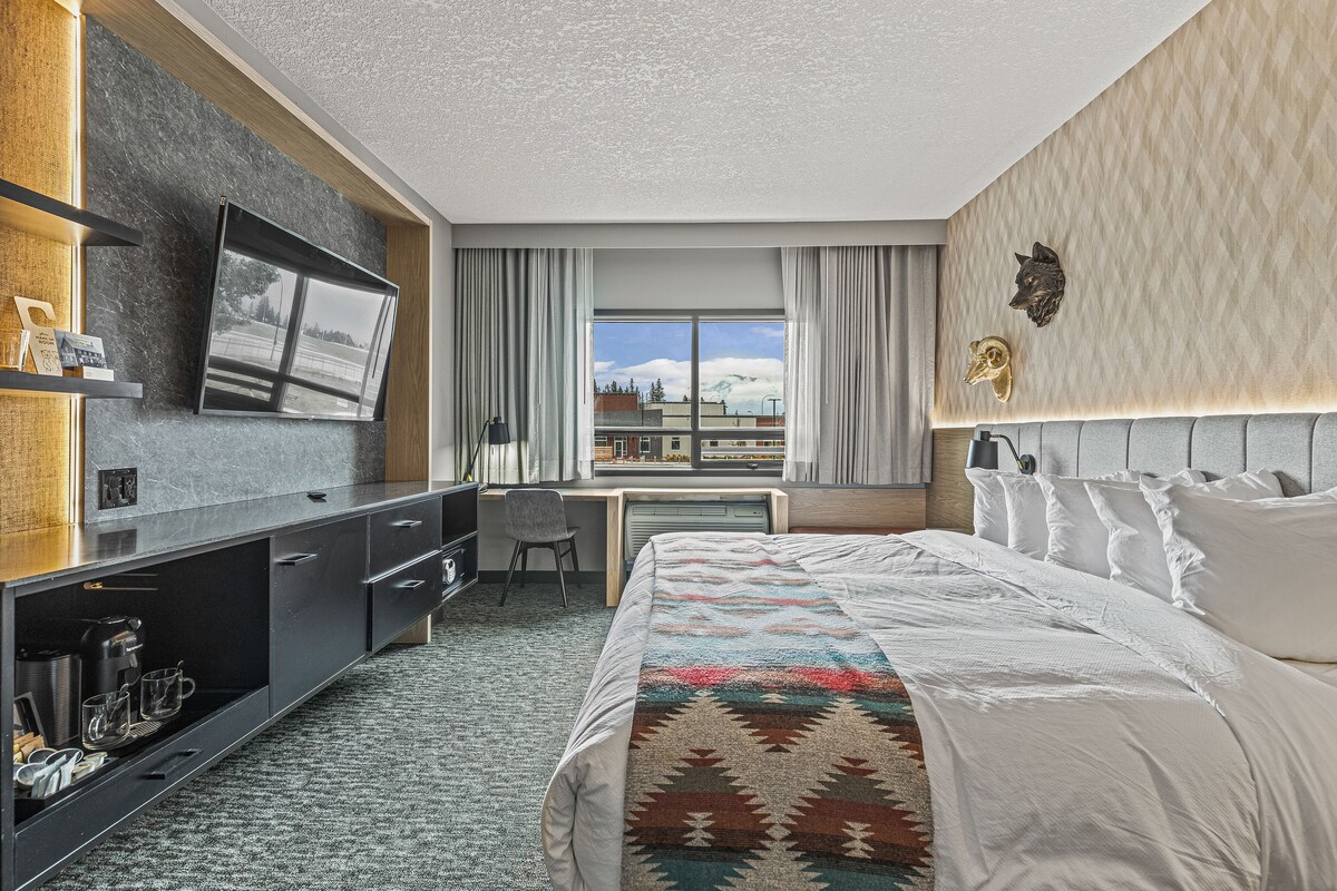 A modern hotel room with a large bed, a desk, a TV, and a view of the city.