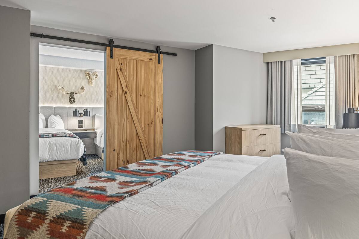 Une chambre d’hôtel moderne avec un grand lit, une porte coulissante en bois, une table de chevet, une petite table et une fenêtre avec des rideaux.