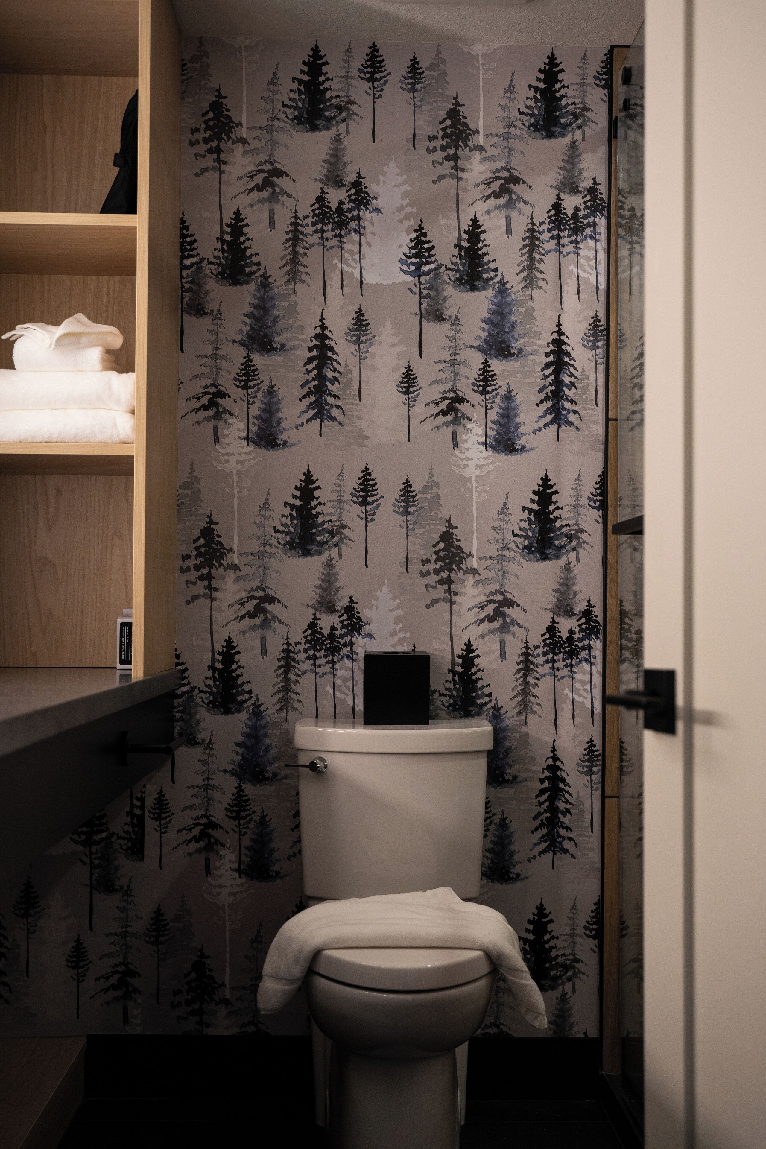 Un bagno con un water, uno scaffale con asciugamani piegati e una parete con motivo foresta.