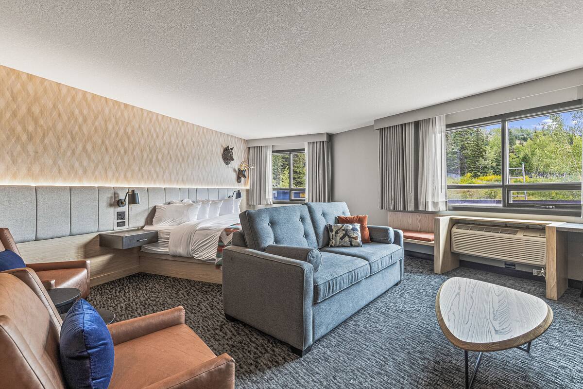 Une chambre d’hôtel avec un canapé, un fauteuil et une petite table. On y trouve un lit avec une tête de lit et une fenêtre avec des rideaux.