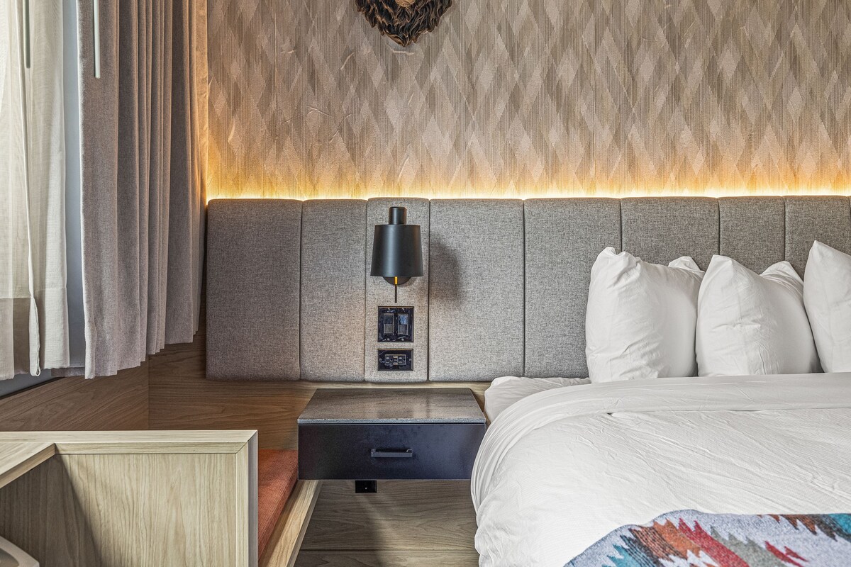 A modern hotel room with a large bed, a bedside table, a nightstand, a wall-mounted lamp, and a textured wall panel.