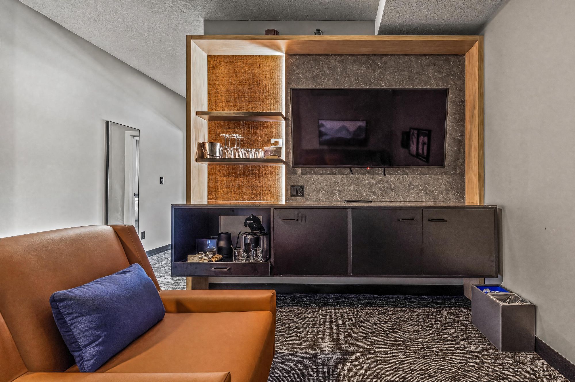 Un salon moderne avec un canapé en cuir, un téléviseur à écran plat fixé à un mur texturé, un appareil à café intégré et un tapis à motifs.