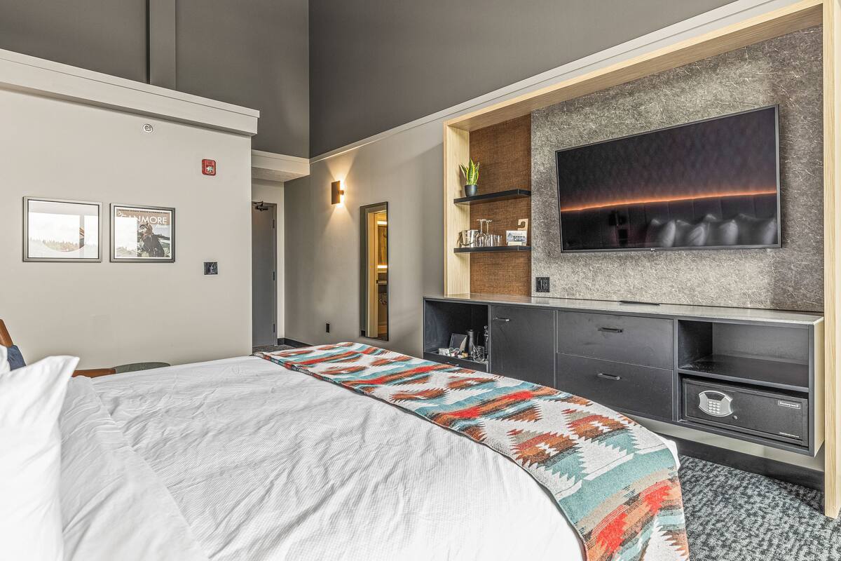 Une chambre d’hôtel avec un lit, un téléviseur fixé à un mur texturé, une petite étagère avec une plante et une porte donnant sur une autre pièce.