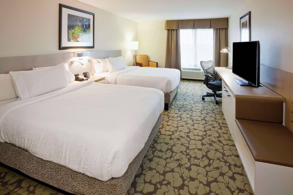 Quarto de hotel com duas camas, uma escrivaninha com computador, uma cadeira e uma televisão.