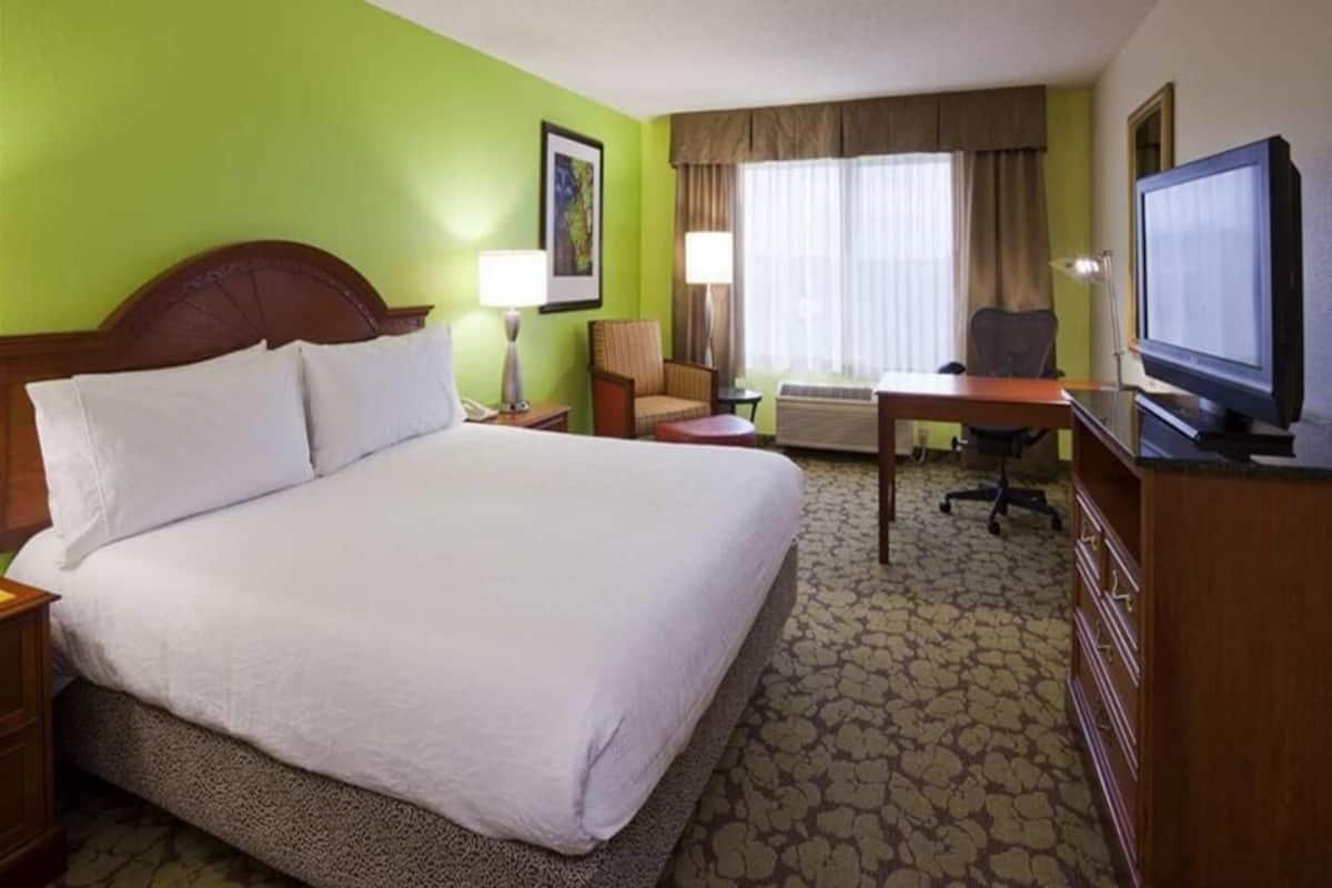 Quarto de hotel com uma cama grande, uma mesa, uma cadeira, uma televisão e uma janela com cortinas.