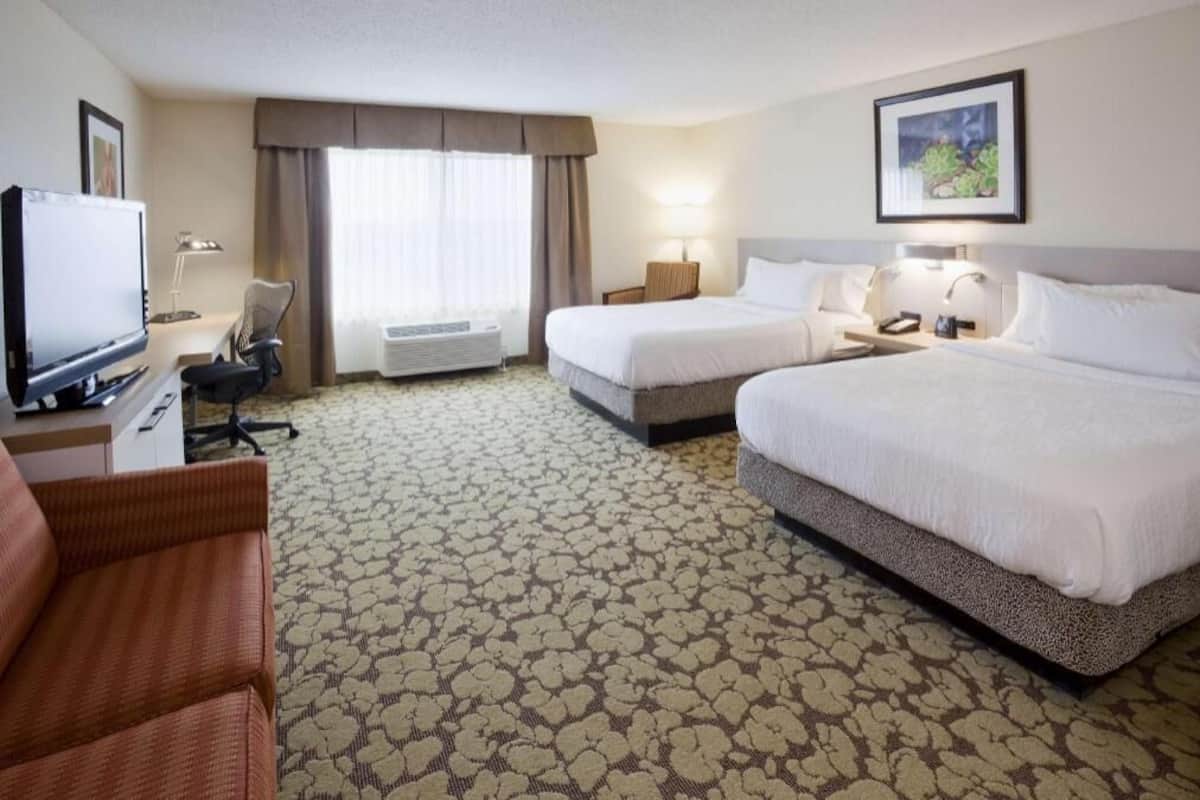 Quarto de hotel com duas camas, uma escrivaninha com computador, uma cadeira, ar condicionado e uma televisão.