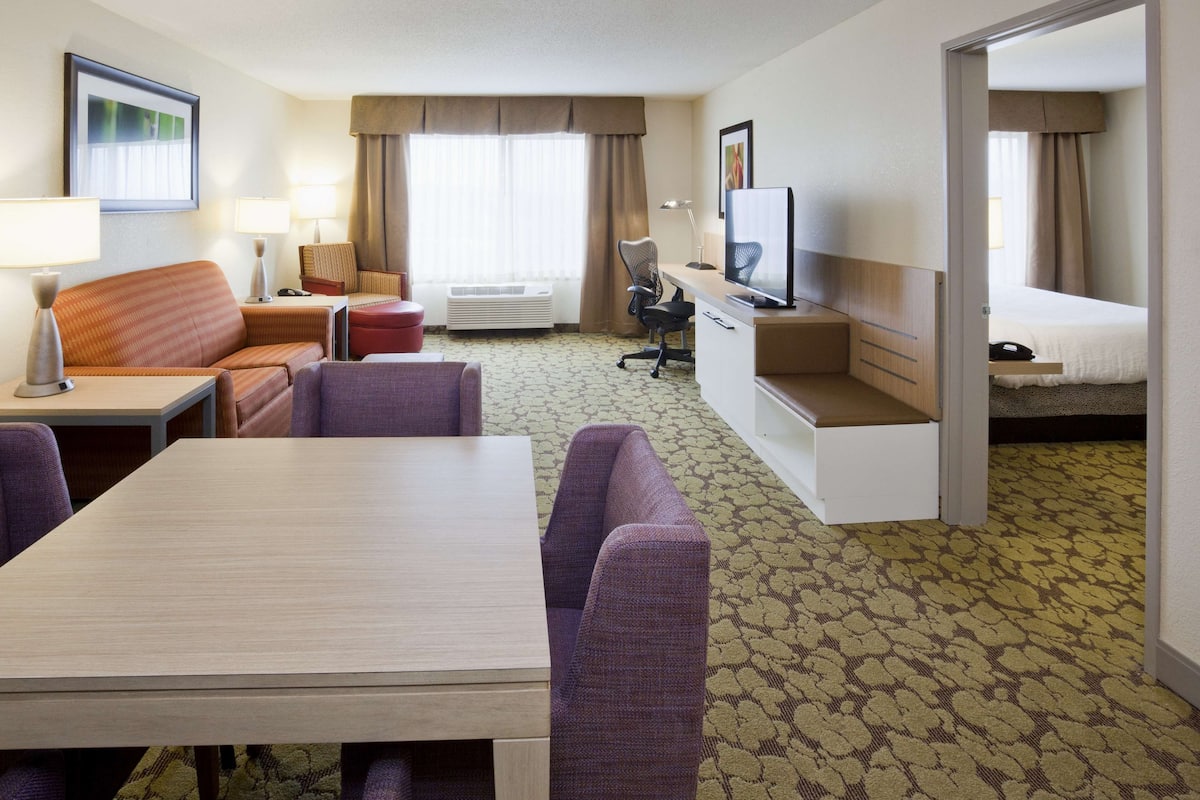Quarto de hotel com cama, escrivaninha, computador, televisão, sofá e mesa de jantar.