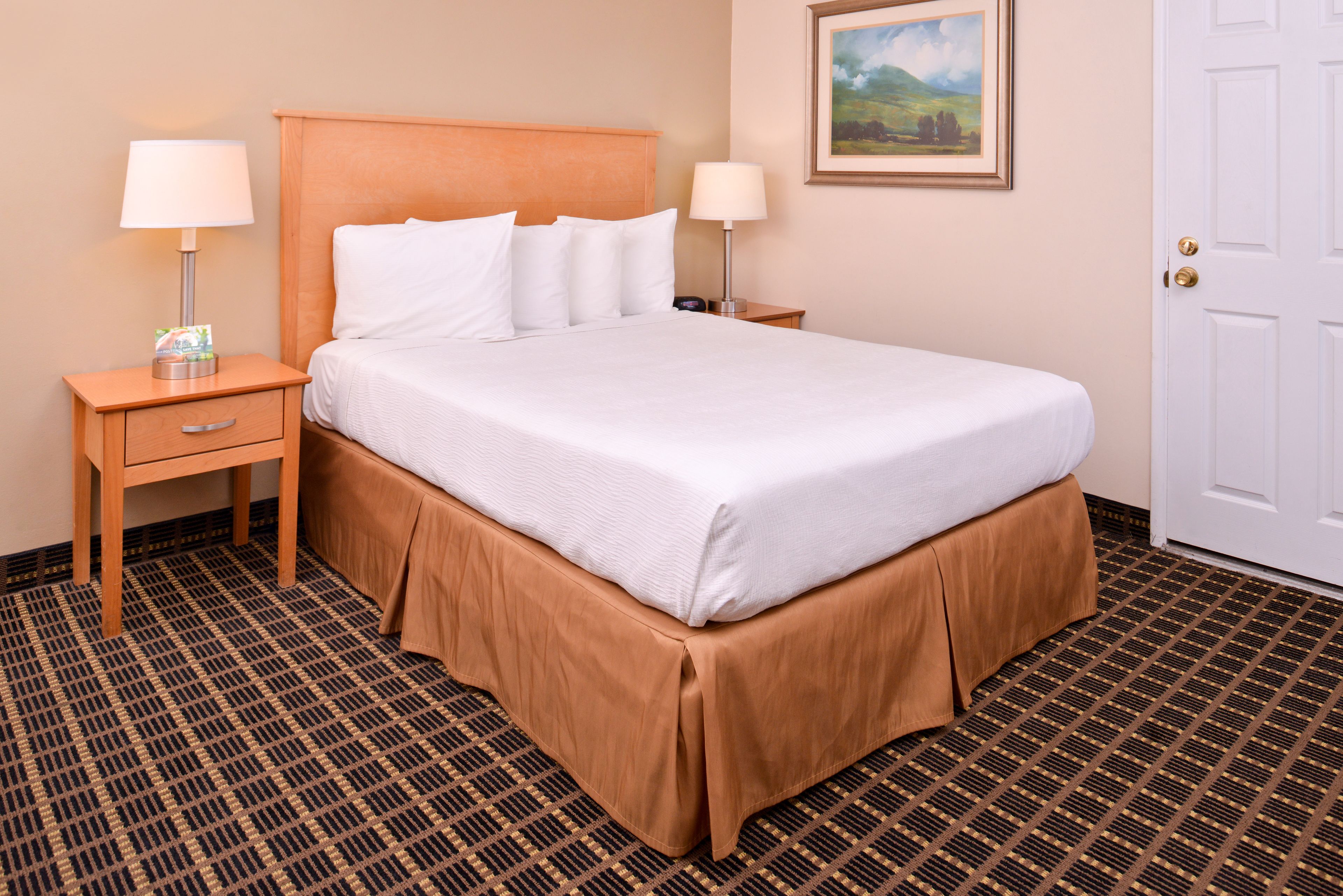 A neatly made bed with white linens, a wooden headboard, and a brown skirt. There are two bedside tables with lamps and a framed landscape painting on the wall.