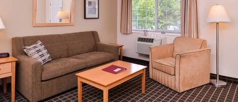 A hotel room with a brown sofa, a wooden coffee table, a beige armchair, a standing lamp, and a window with curtains.