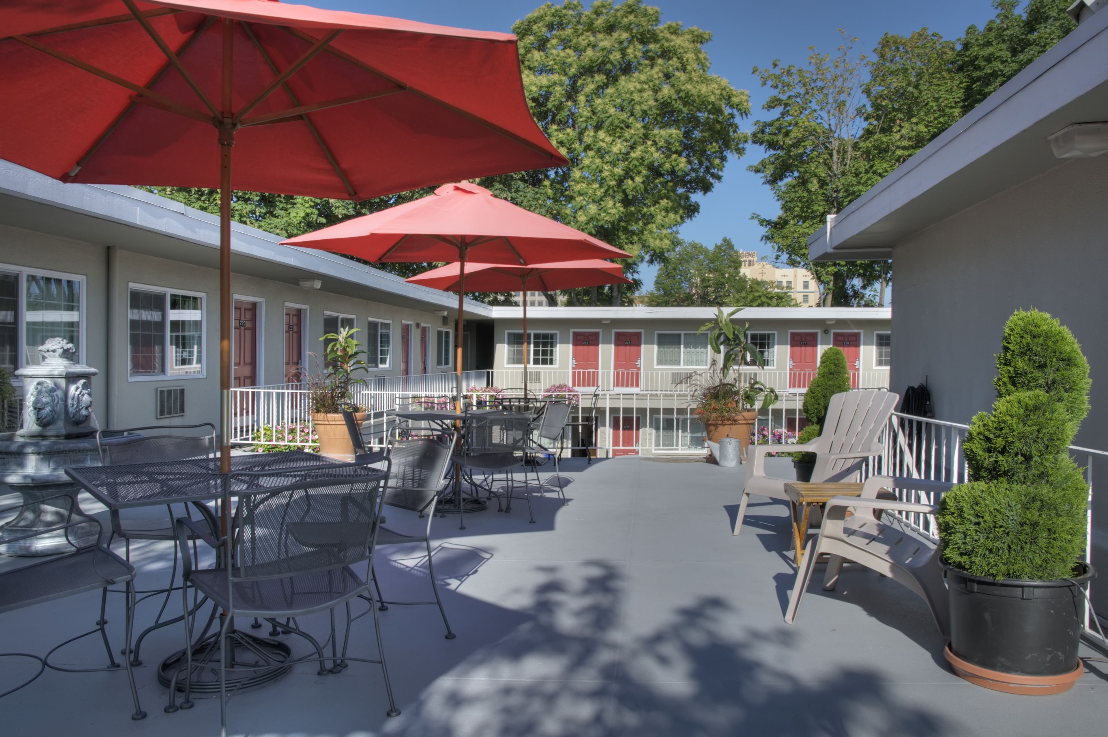 Eine überdachte Terrasse im Freien mit Metalltischen und -stühlen, roten Sonnenschirmen und Kübelpflanzen.