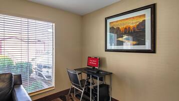 A room with a desk, chair, and a computer. There is a window with blinds, a framed picture on the wall, and a carpeted floor.