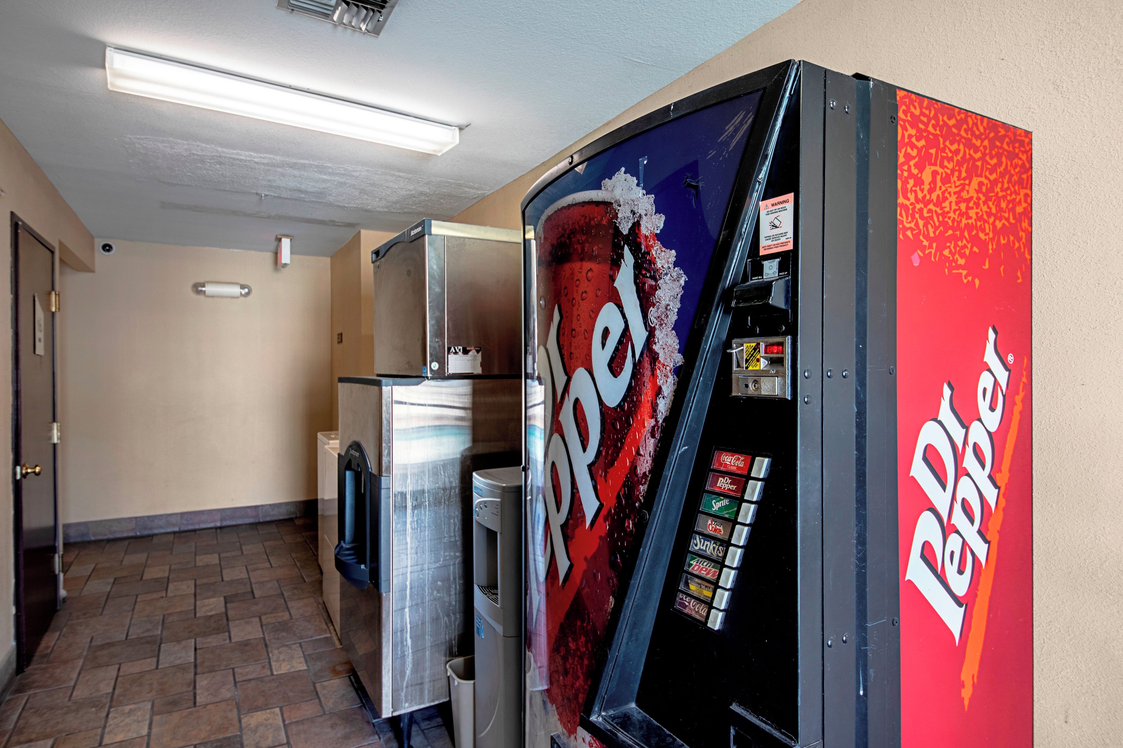 Una máquina expendedora con el logotipo de Dr Pepper, ubicada en un pasillo con piso de baldosas y una puerta a la izquierda.
