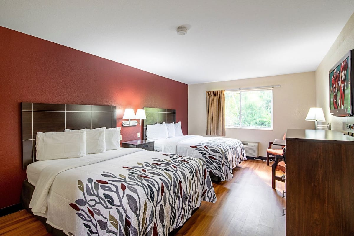 Une chambre d’hôtel avec deux lits, un bureau, une chaise, une fenêtre avec des rideaux et un tableau au mur.