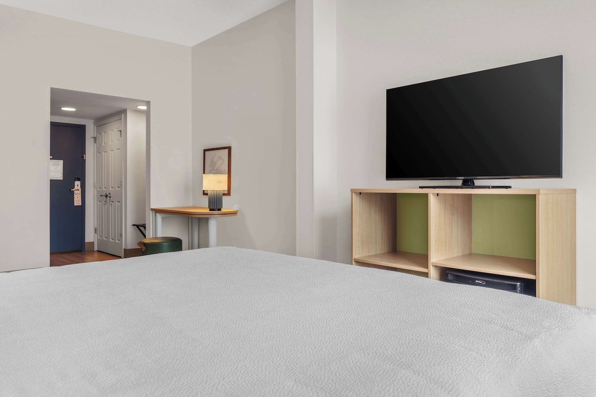 A hotel room with a large bed, a flat-screen TV mounted on the wall, a small table with a lamp, and a door leading to another room.