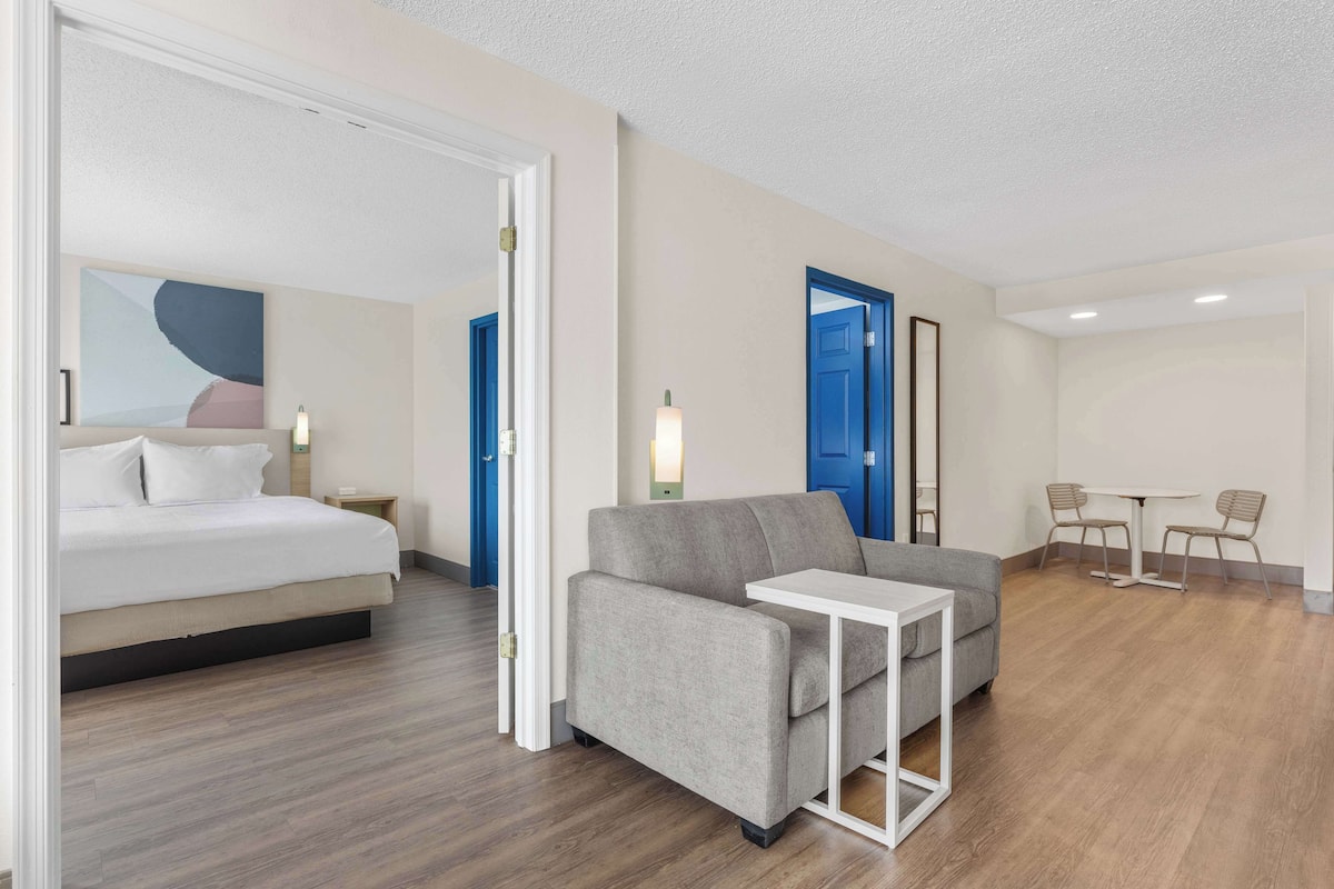 A modern hotel room with a bed, sofa, and a small table. There are two doors, one blue and one white, and a small table with chairs.