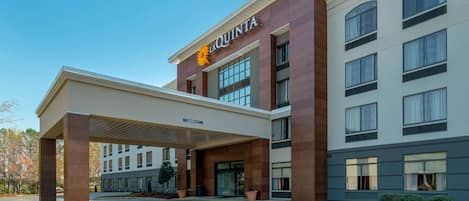Un hotel moderno con un llamativo letrero de 'La Quinta', estacionamiento techado y un área de entrada ajardinada.
