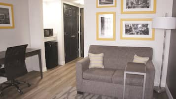 A compact living space with a grey sofa, a small side table, a black office chair, a microwave, and framed pictures on the wall.