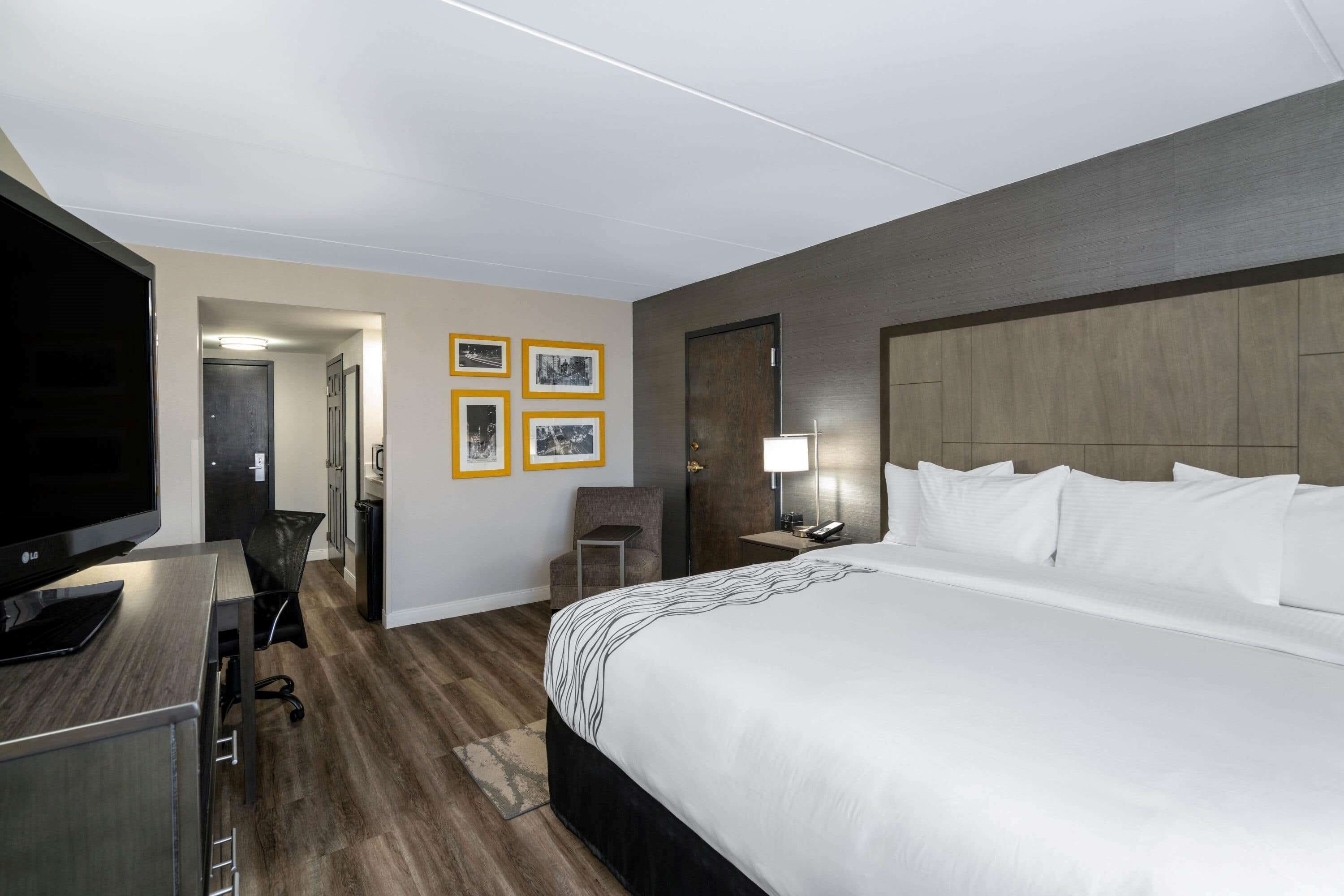 A hotel room with a large bed, a TV, a desk, and a chair. There are framed pictures on the wall and a lamp on the bedside table.