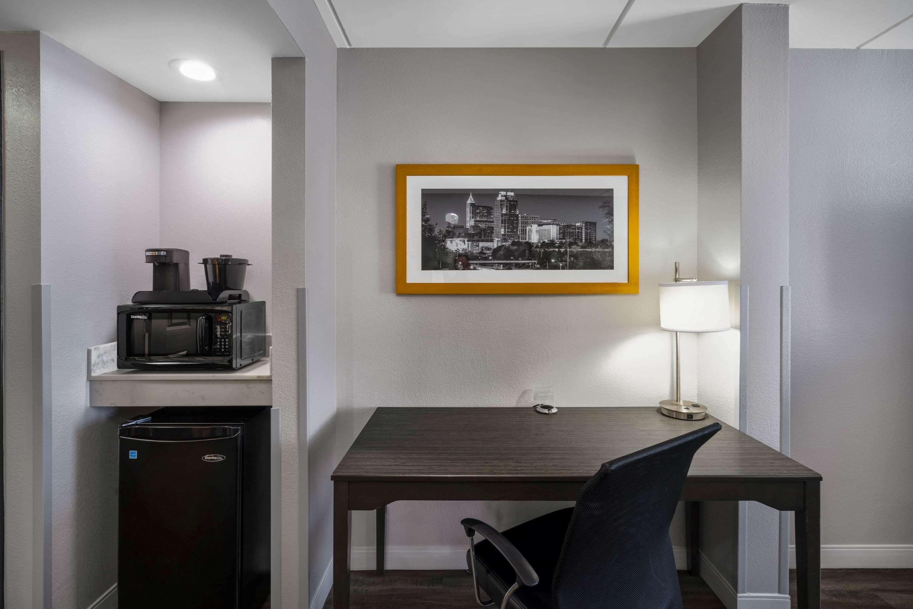 A compact hotel room with a kitchenette, a dining table with a chair, a framed cityscape picture on the wall, and a bedside lamp.