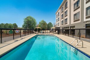 A swimming pool with lounge chairs and umbrellas, surrounded by a fence and a building with multiple windows.