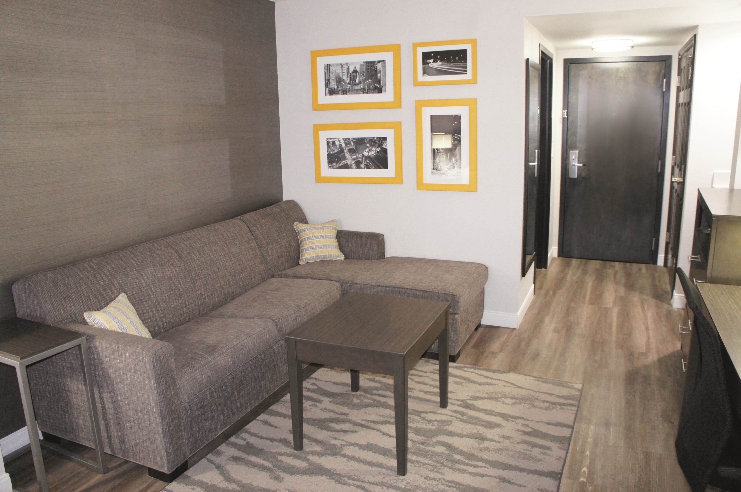 A living room with a sectional sofa, a coffee table, and a rug. There are framed pictures on the wall and a door in the background.