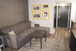 A living room with a sectional sofa, a coffee table, and a rug. There are framed pictures on the wall and a door in the background.