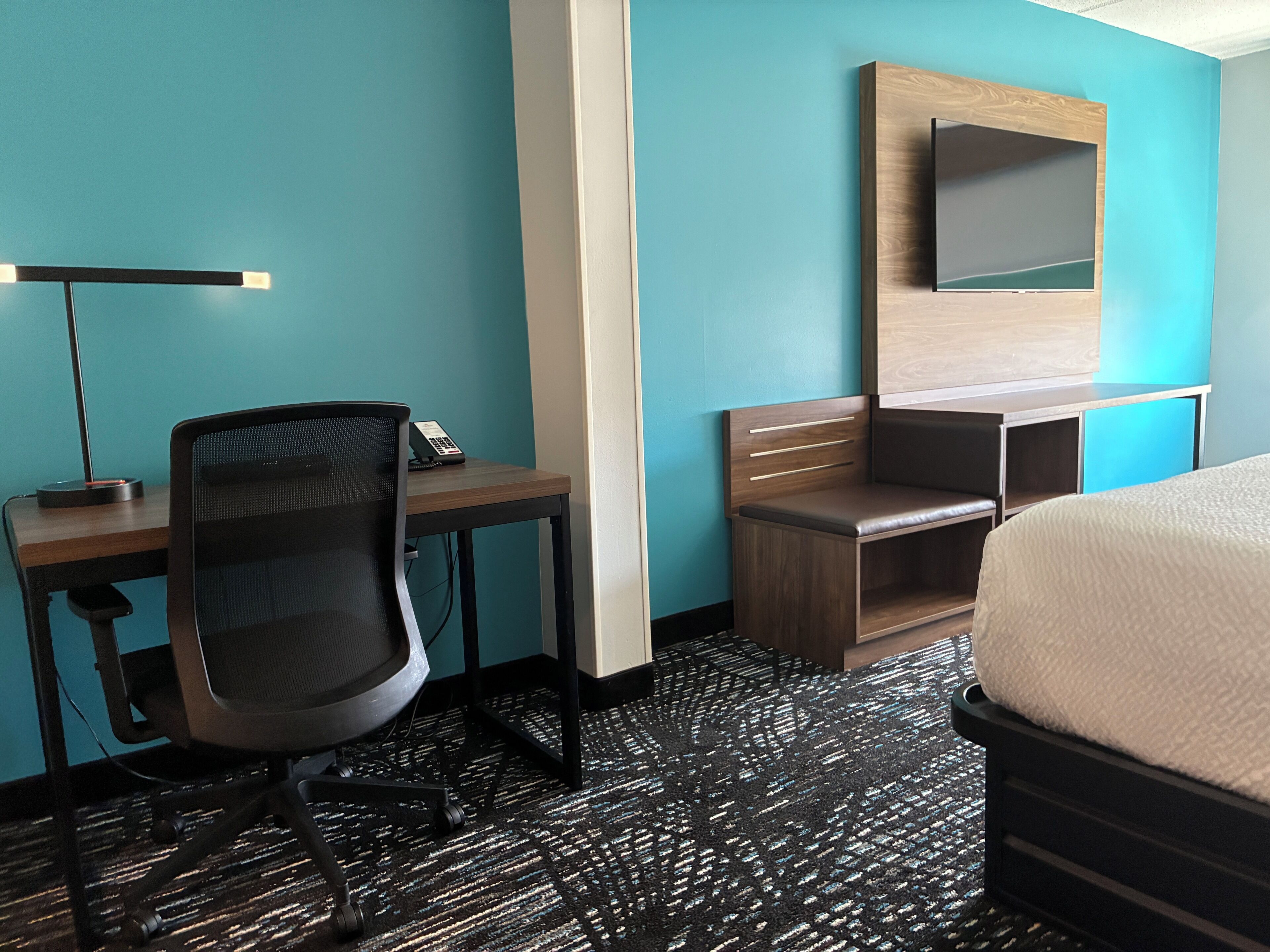 Une chambre d’hôtel équipée d’un bureau, d’une chaise, d’un lit et d’un téléviseur fixé au mur.