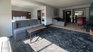 Une chambre d’hôtel moderne avec un canapé gris, une petite table, un tapis à motifs, un lit avec du linge de lit blanc, un bureau avec un ordinateur et un miroir.