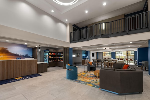 A spacious lobby with a reception desk, a seating area with a sofa and chairs, a staircase, and a dining area with tables and chairs.