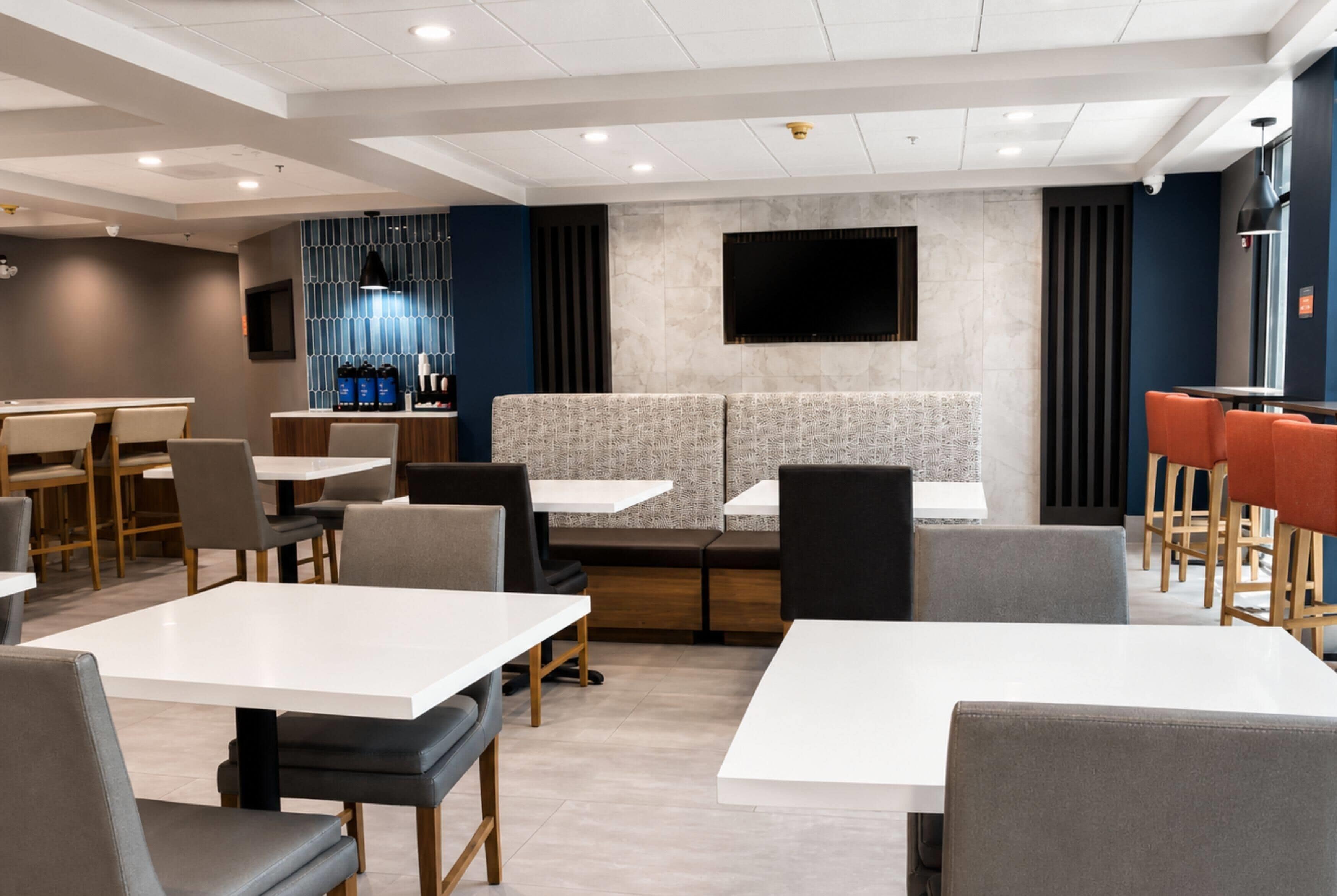 Un restaurant moderne avec des tables blanches et des banquettes grises, un téléviseur à écran plat fixé au mur et un coin café.