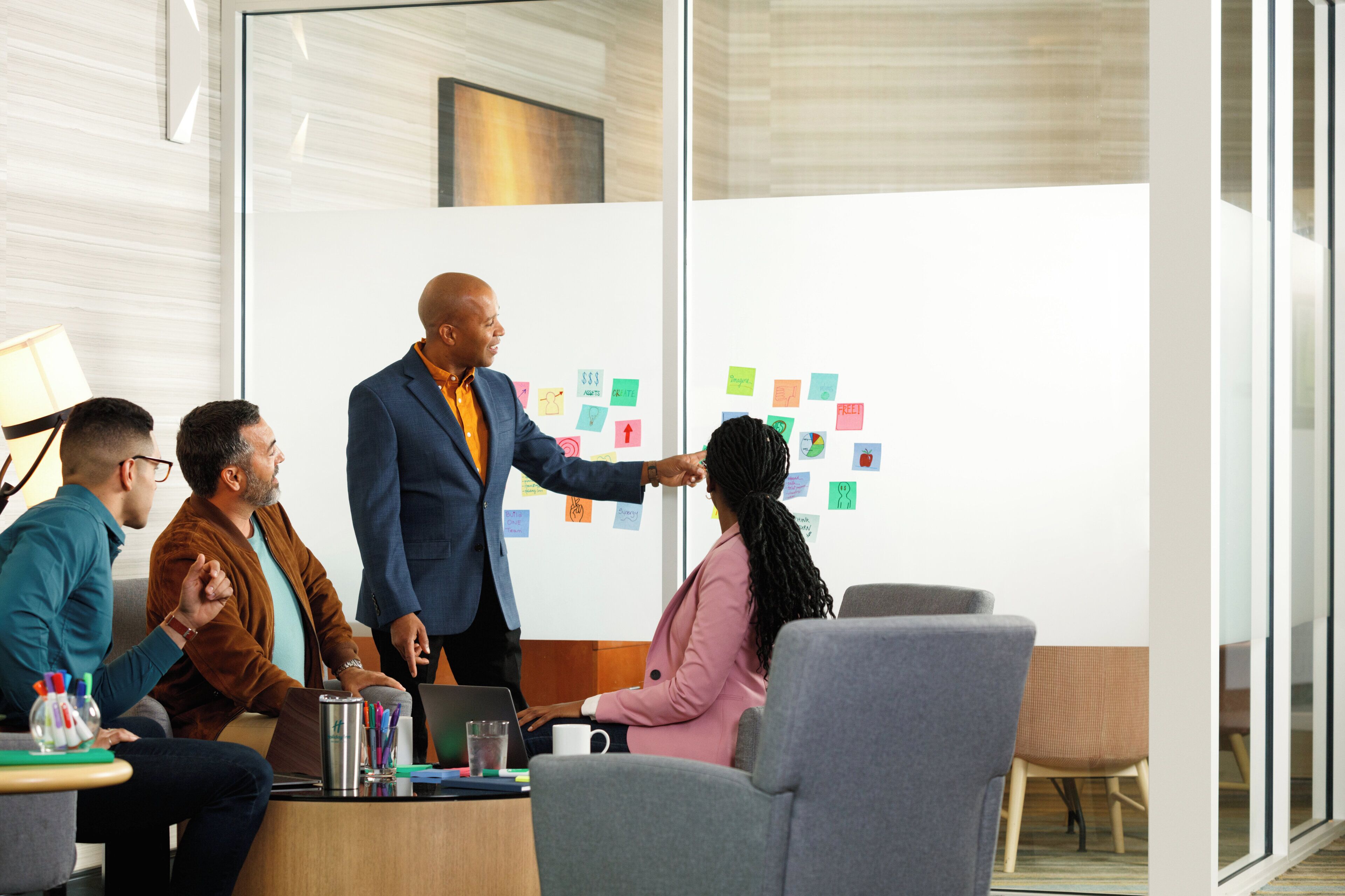 A meeting room with a glass wall, a whiteboard with sticky notes, and a table with laptops and pens.