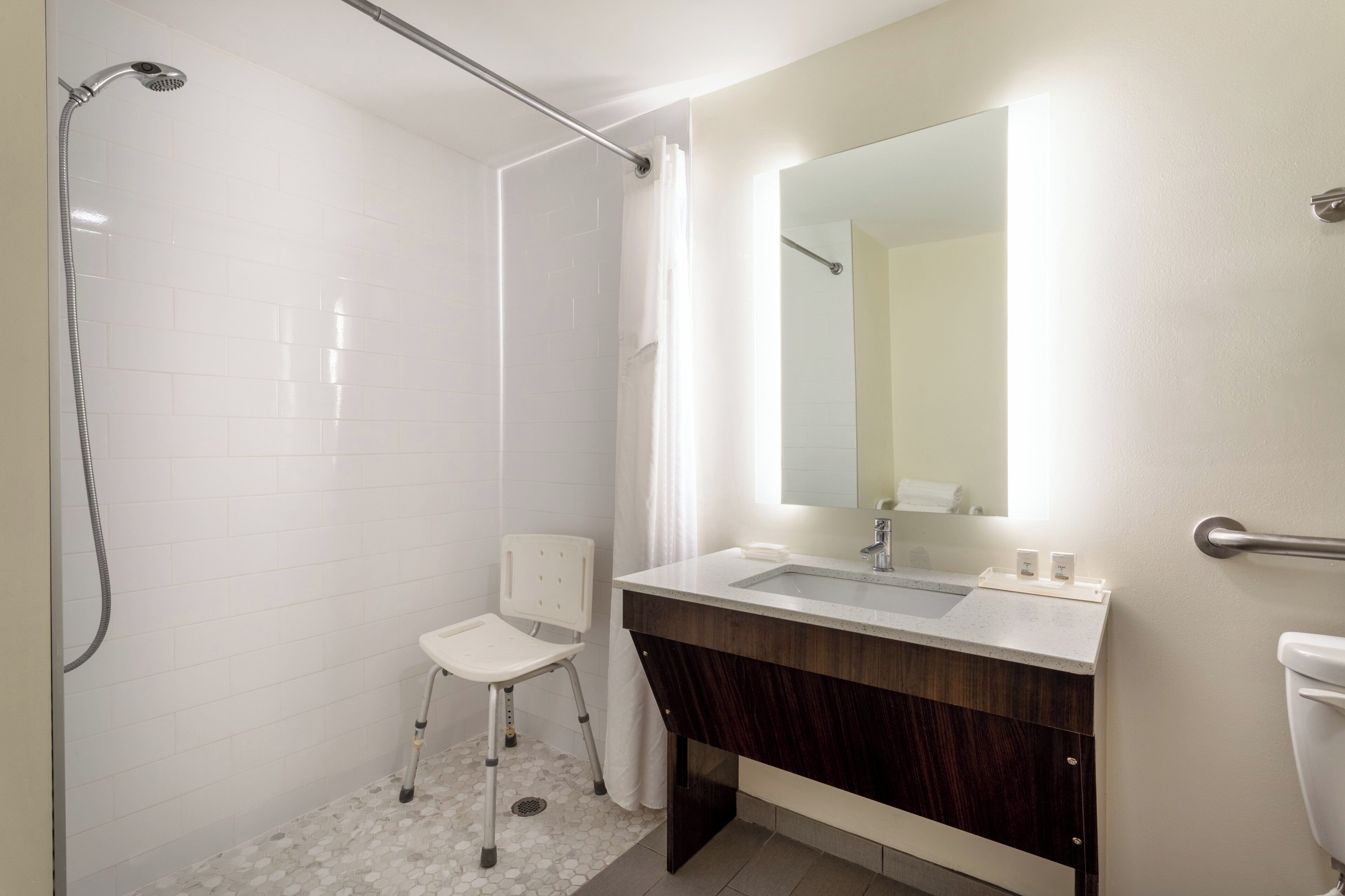A bathroom with a shower, a toilet, a sink with a mirror, and a chair.