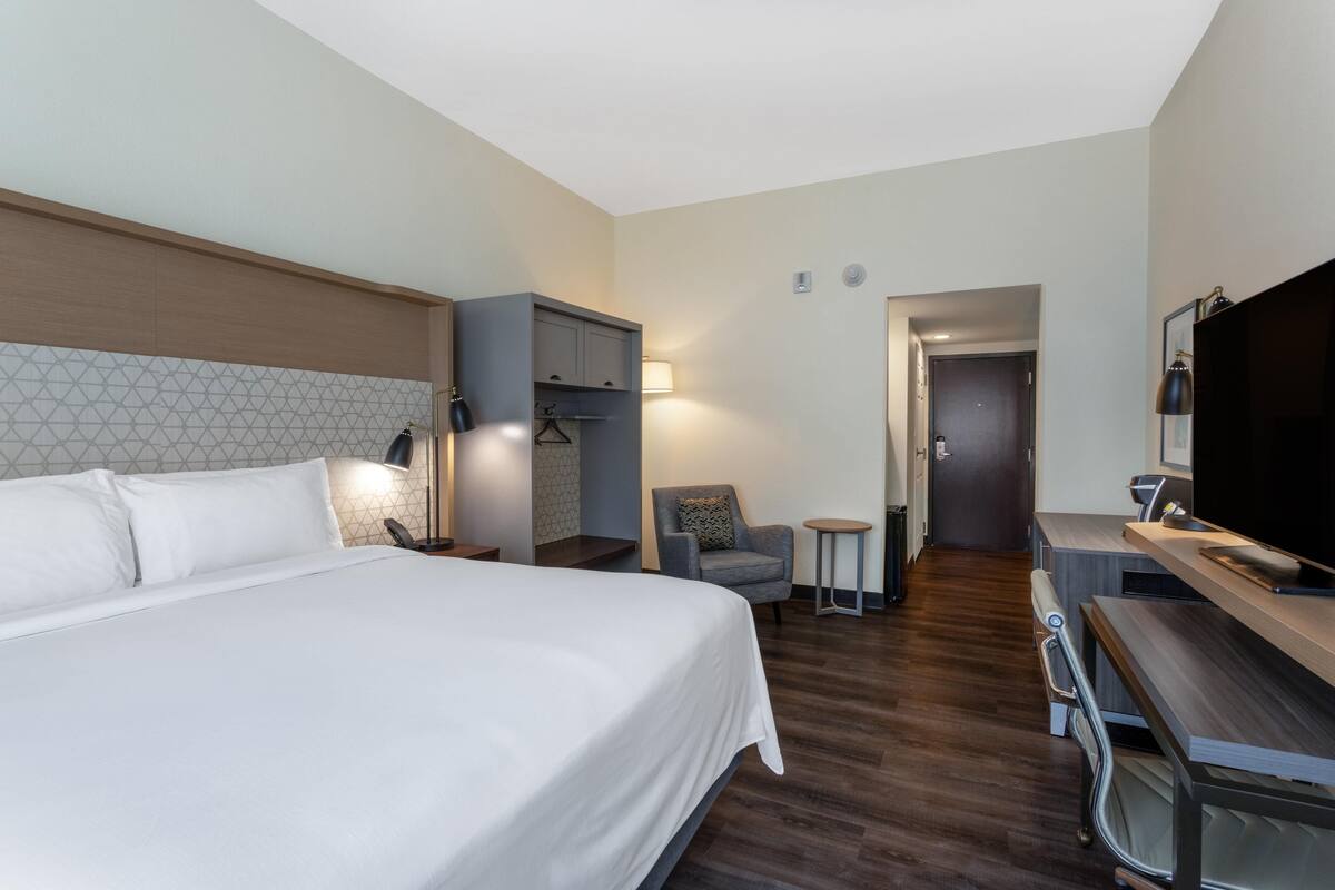 A hotel room with a large bed, a desk with a flat-screen TV, a chair, a small table, and a wardrobe.