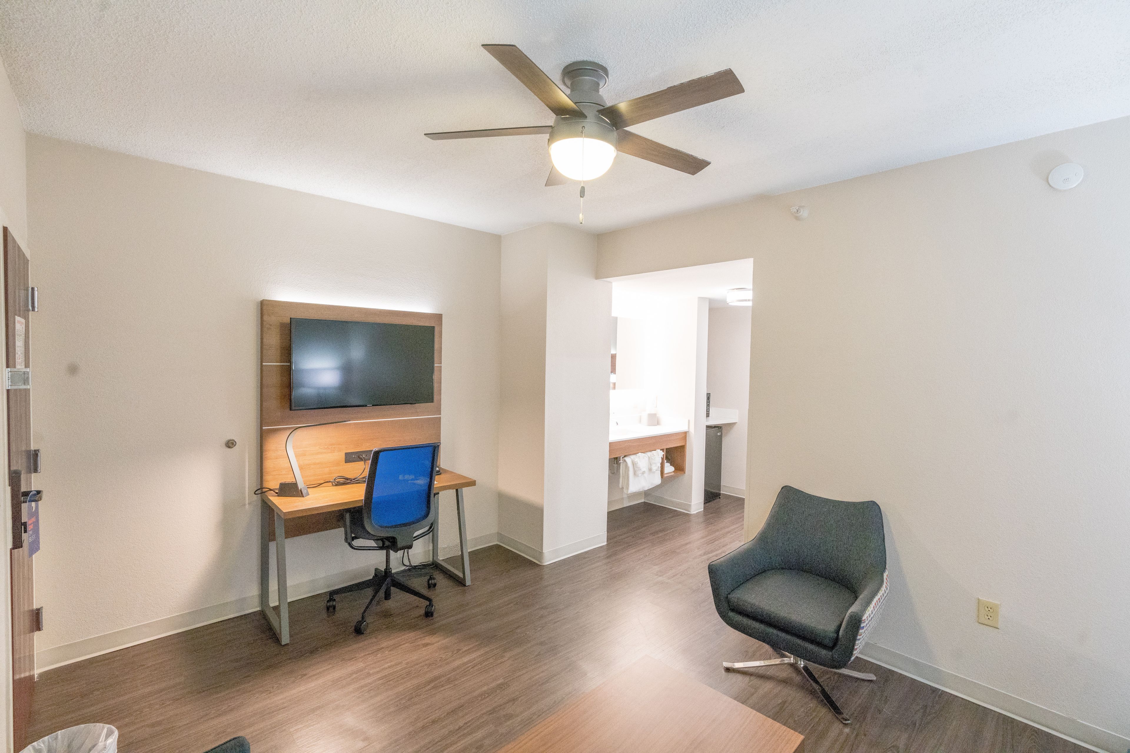 Een moderne woonkamer met een houten vloer, een plafondventilator, een flatscreen televisie aan de muur, een bureau met stoel en een grijze fauteuil.