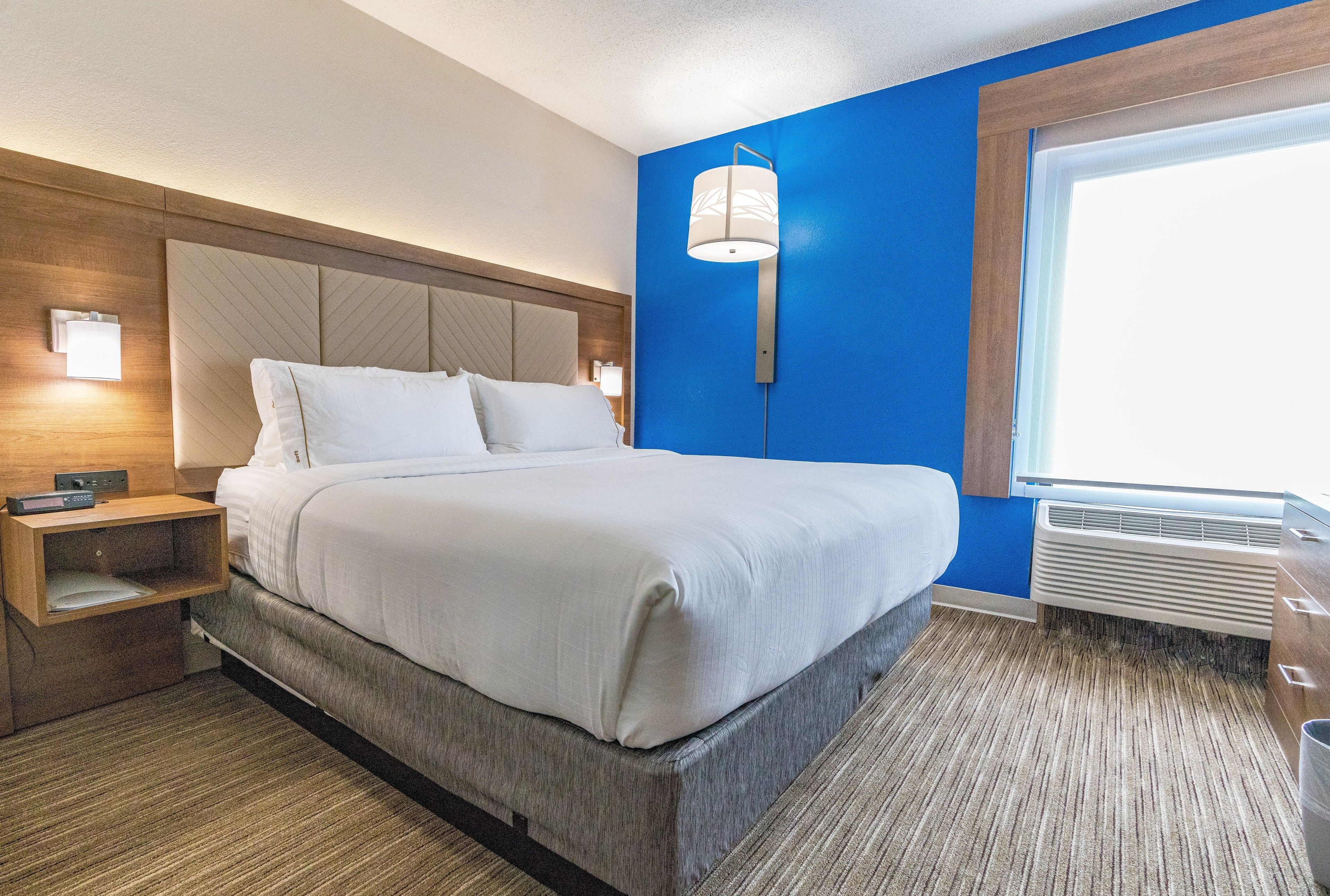 A hotel room with a bed, bedside tables, a wall-mounted lamp, and a window with blinds.