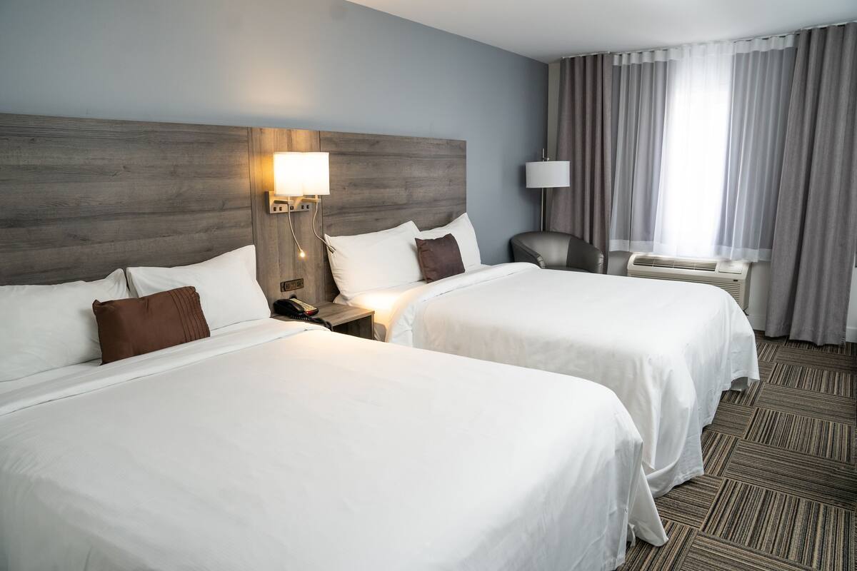 Une chambre d’hôtel avec deux lits, une table de chevet avec une lampe, un bureau avec une chaise et une grande fenêtre avec des rideaux.