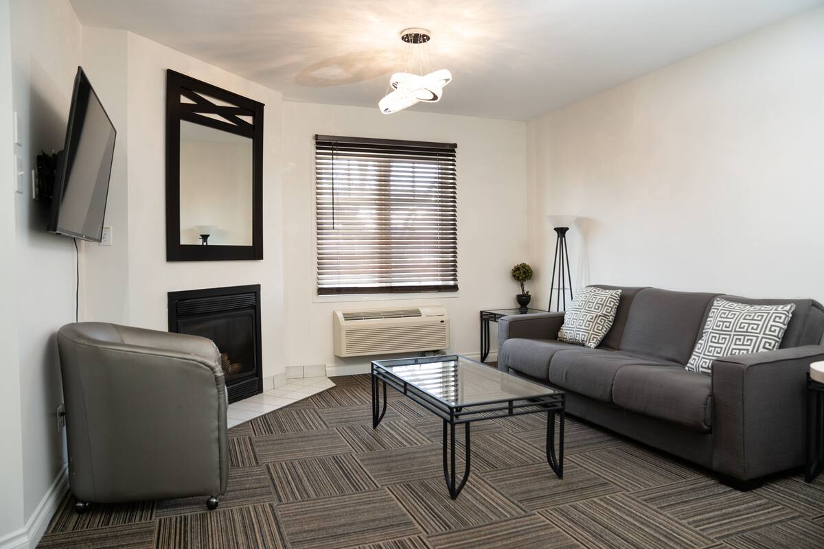 Un salon moderne avec un canapé, un fauteuil et une table basse en verre. On y trouve une cheminée, un grand miroir et un luminaire suspendu au plafond.