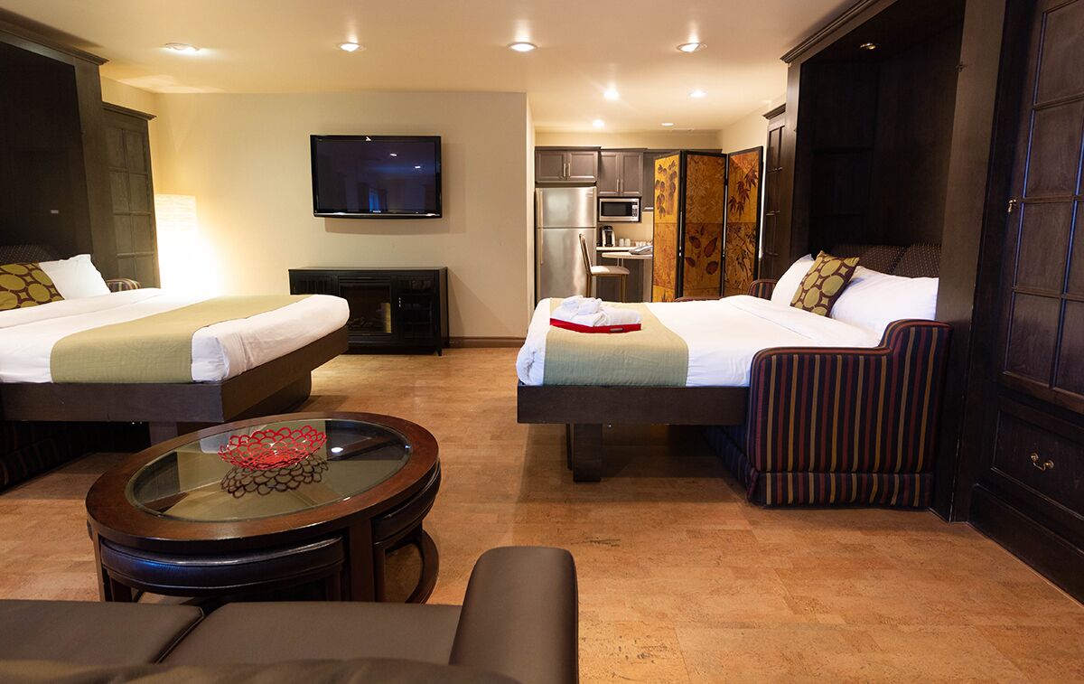A hotel room with two beds, a TV, a kitchen area, and a seating area with a glass table.