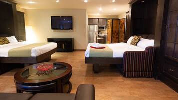 A hotel room with two beds, a TV, a kitchen area, and a seating area with a glass table.